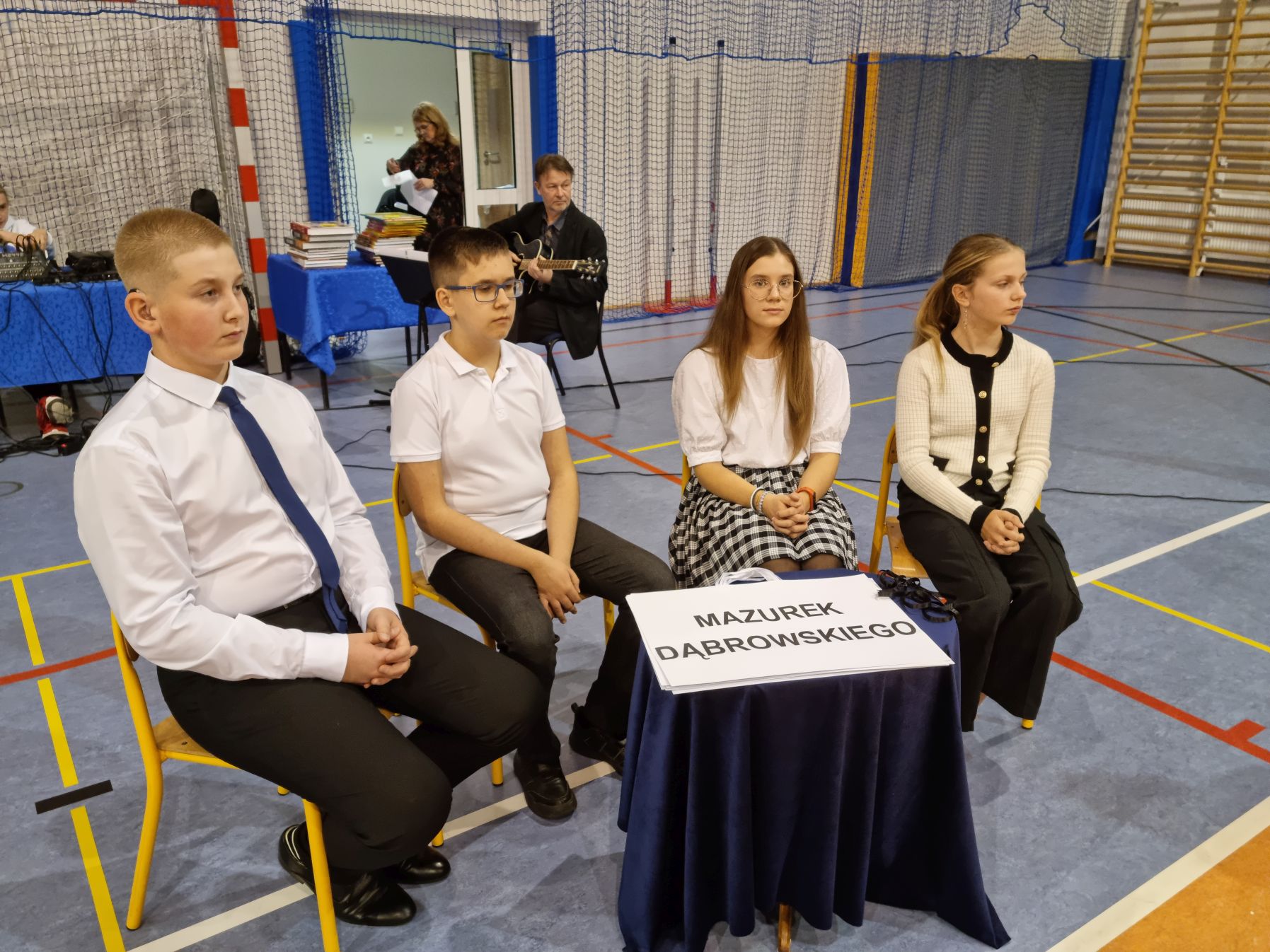 Czworo dzieci ubranych w czarno-białe stroje siedzi przy stoliku, w tle nauczyciel z gitarą