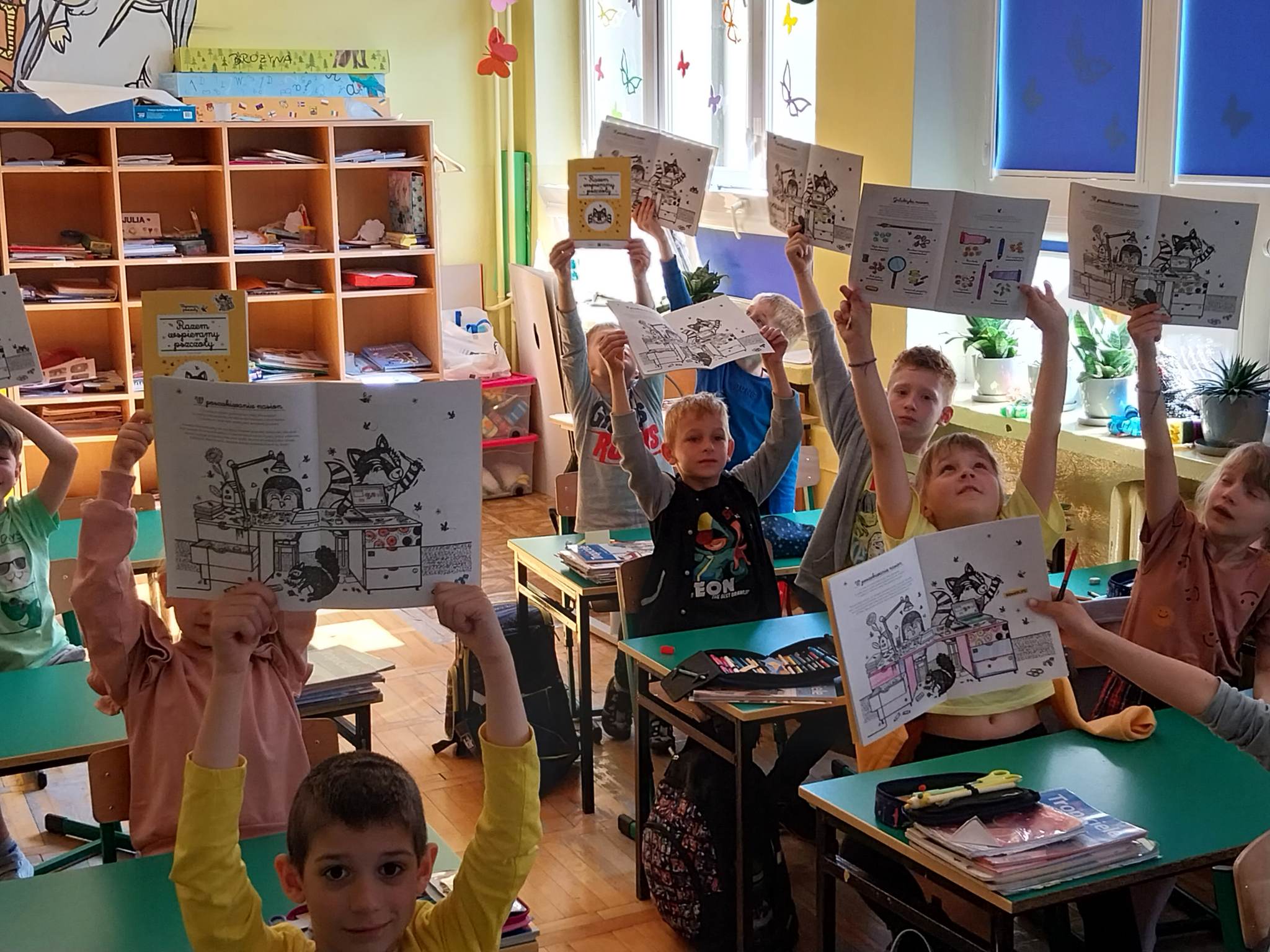 Uczniowie w ławkach z podniesionymi zeszytami ćwiczeń
