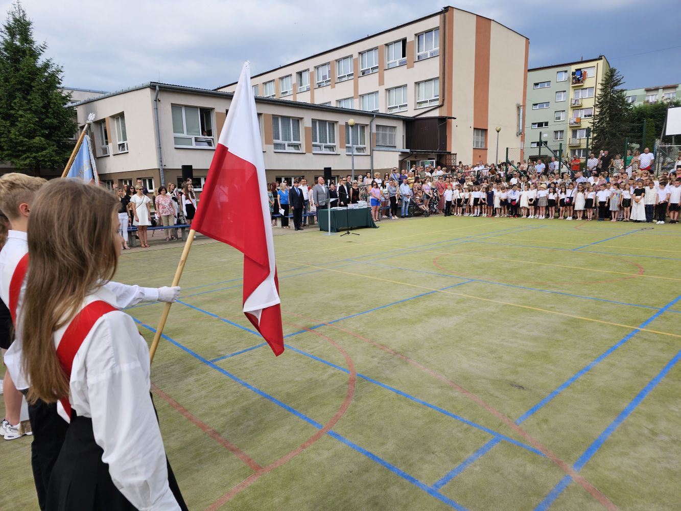 Boisko szkolne, poczet sztandarowy, w głębi uczniowie, dyrekcja, nauczyciele